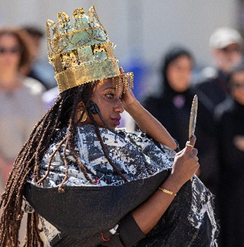 A performer in an elaborate gold crown and patterned robe, holding a blade, in a crowd.