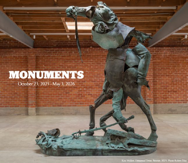A large bronze sculpture of entangled figures, installed in a brick-walled gallery beneath the word MONUMENTS.