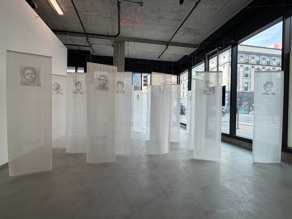 Gallery interior at TnT Art Lab with tall translucent hanging panels, each carrying a printed portrait.